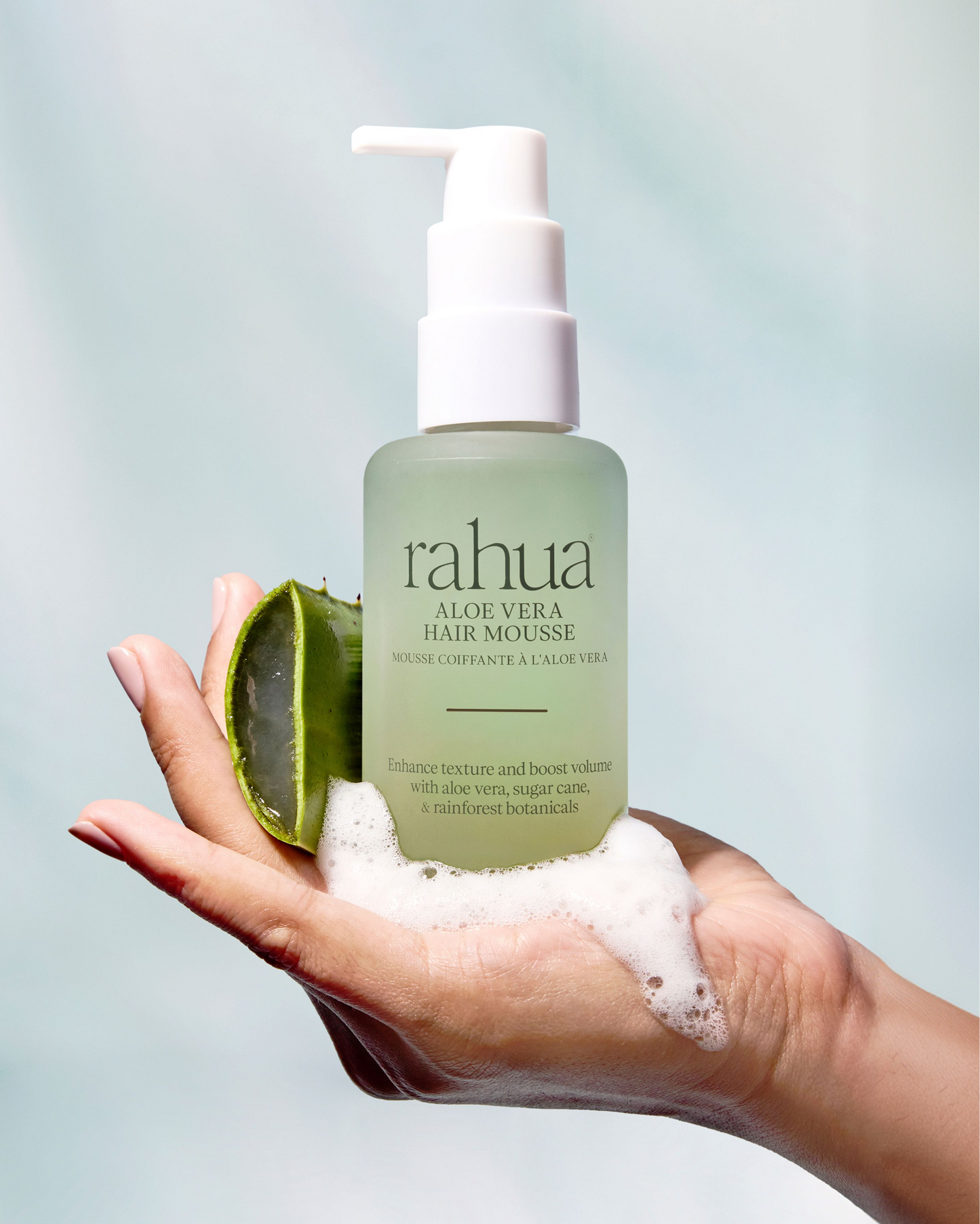 Hand holding a bottle of Rahua Aloe Vera Hair Mousse with aloe vera leaf and foam on a light blue background