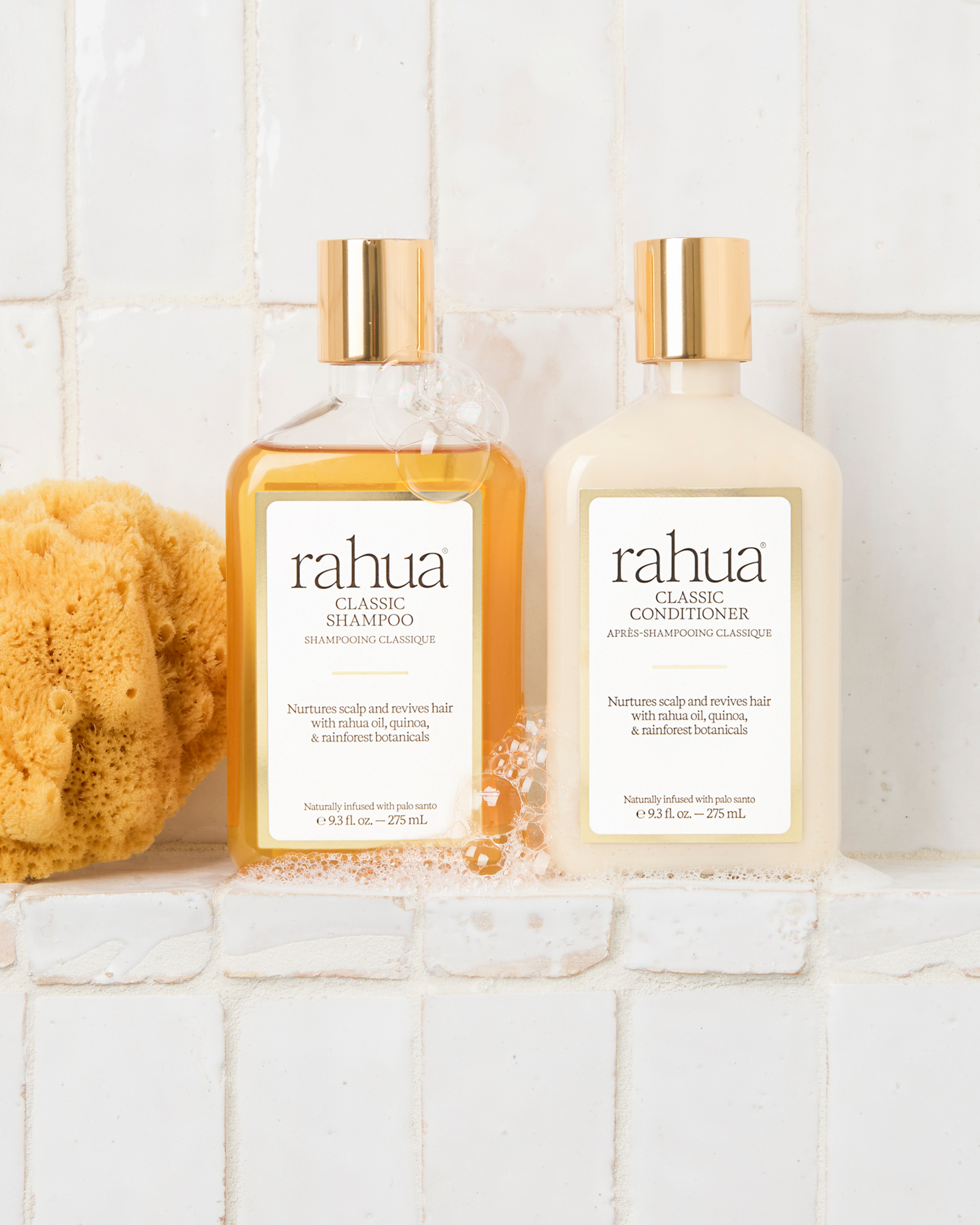 Two bottles of soapy rahua classic shampoo and conditioner on a white tiled shelf with a sponge|variant:93-oz