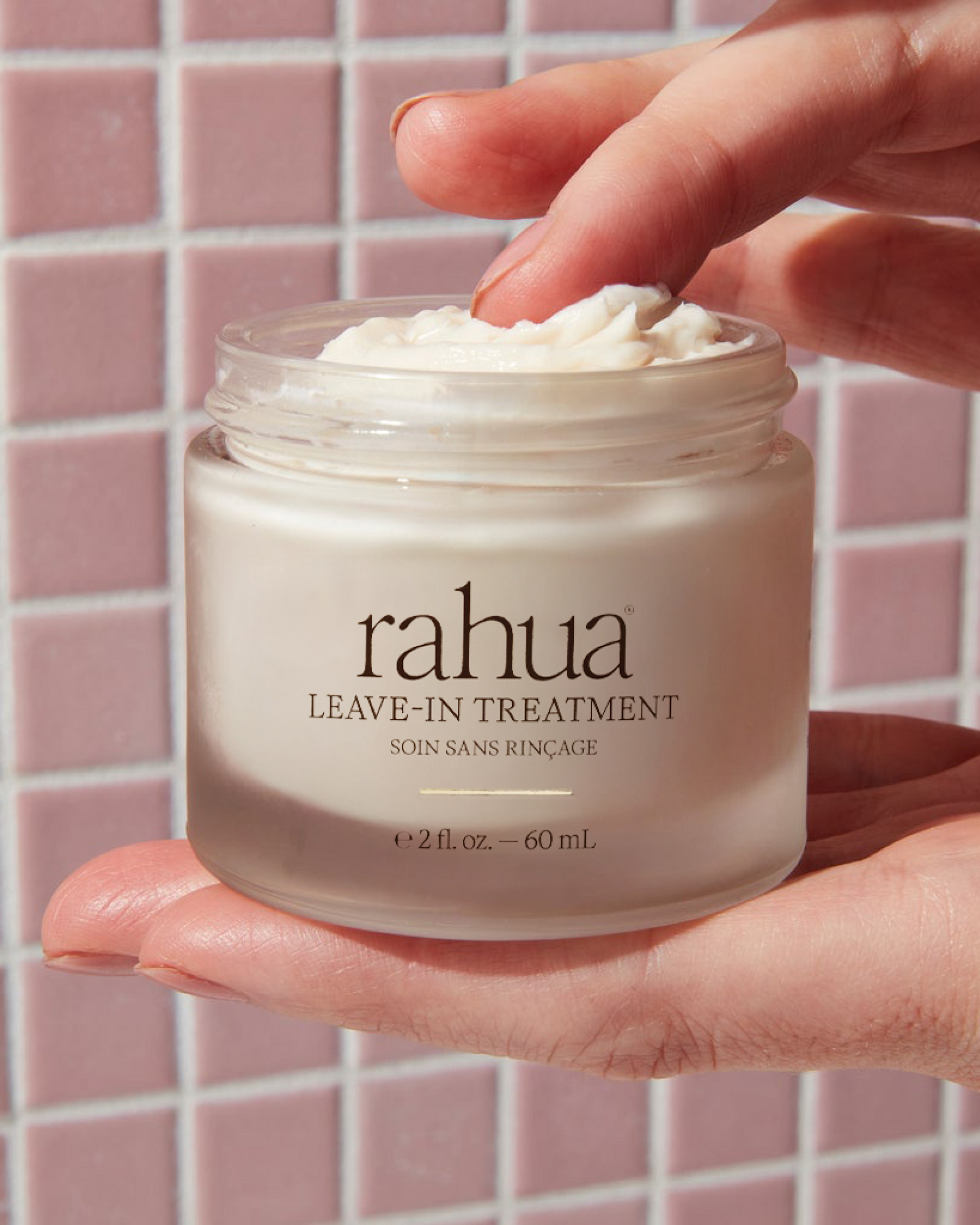 Hand holding a jar of Rahua leave-in treatment against a pink tiled wall.