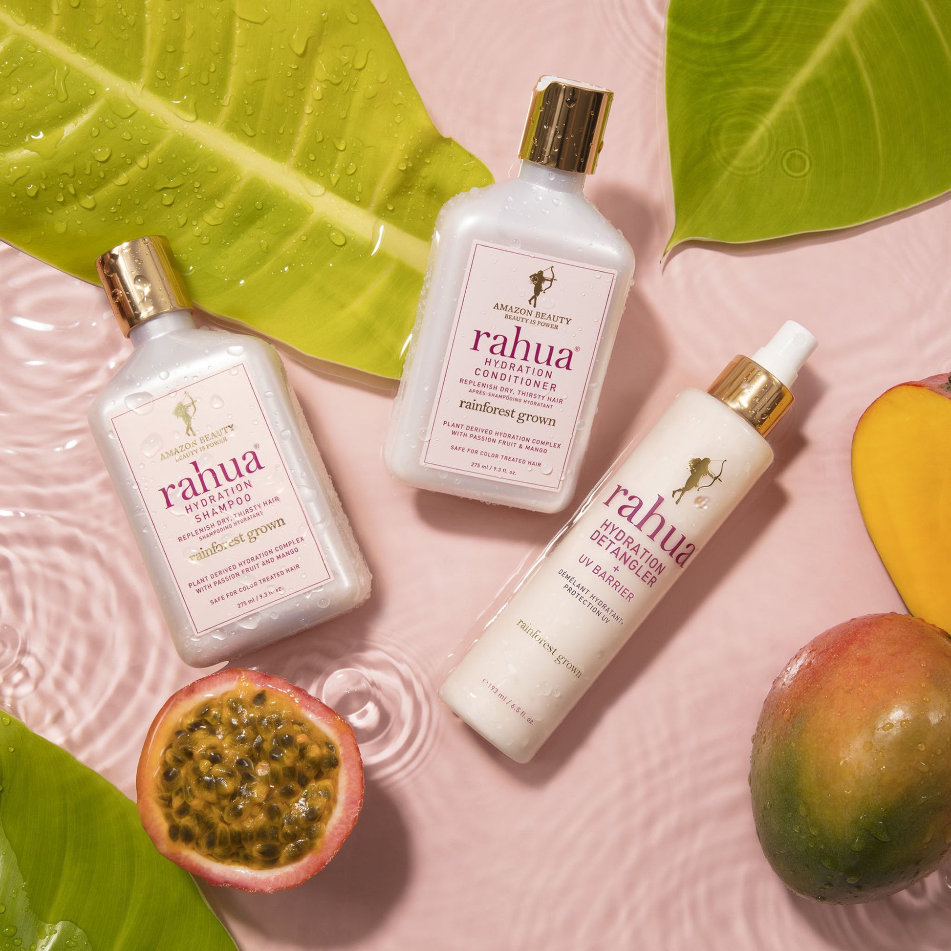 three bottles from rahua's hydration line including hydration shampoo hydration conditioner and hydration detangler on a water submerged pink background with leaves, mangoes and passion fruit|variant:travel-size