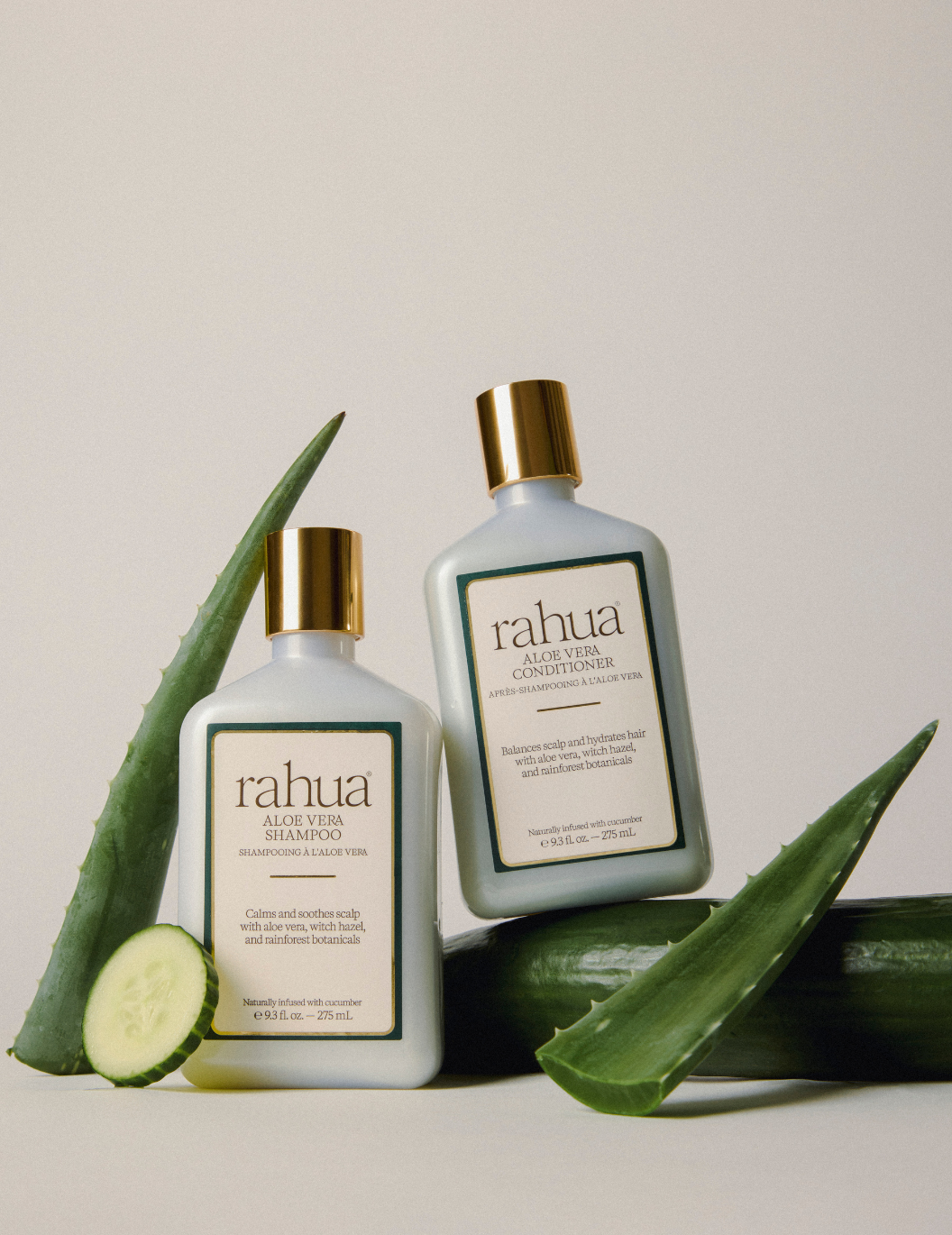 Two bottles of Rahua Aloe Vera Shampoo and Conditioner with aloe vera leaves and a cucumber on a light background