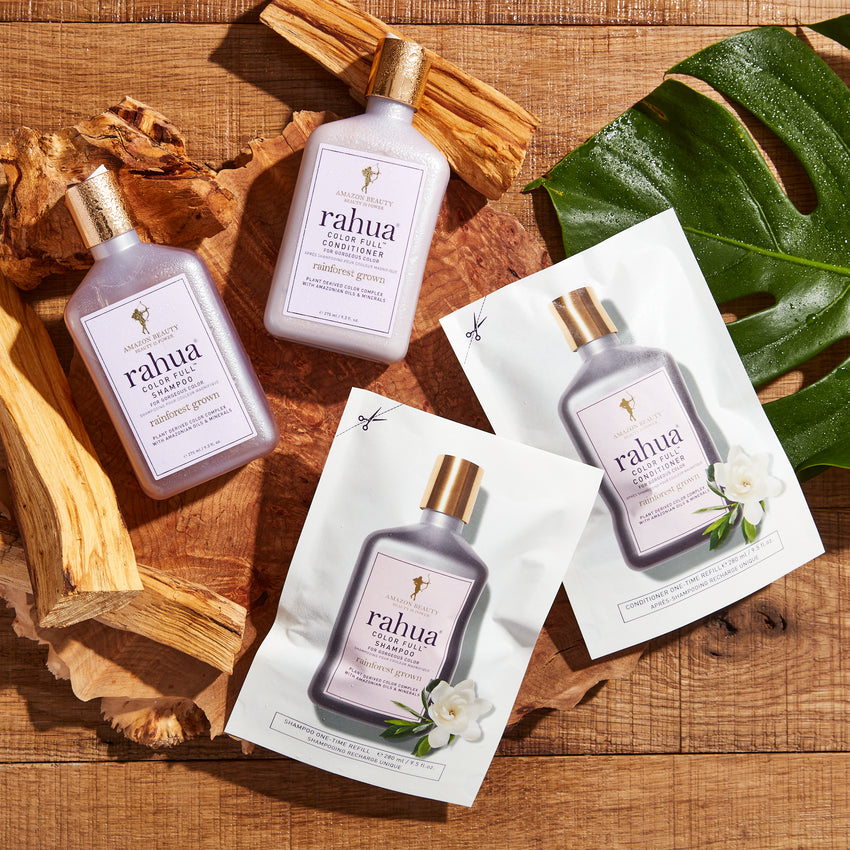 two pouches and two bottles of rahua color full conditioner and shampoo on a wooded background with a palm leaf and wooden pieces