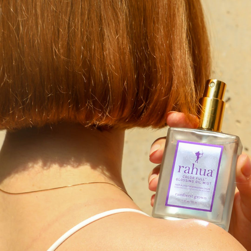 woman holding a bottle of Rahua Color Full Glossing Oil Mist with a white label and blue liquid visible inside. The bottle is designed with a gold-colored cap and a clear glass body