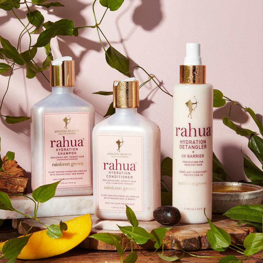 three bottles from rahua's hydration line including hydration shampoo hydration conditioner and hydration detangler on a pink background with leaves, mangoes and rahua oil|variant:full-size