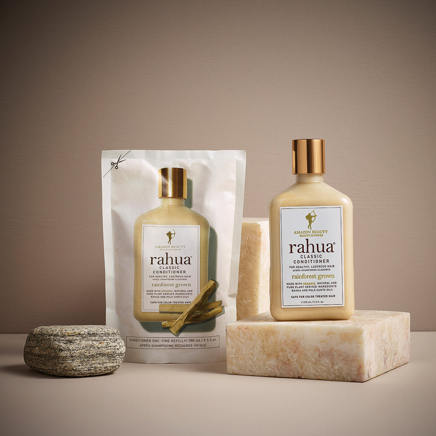 A pouch containing a bottle of Classic Conditioner with a white label, gold cap, and the brand name ' Rahua ' written on it. The product is designed for hair care and is presented in an earth-friendly packaging. A bottle of Classic Conditioner with a gold cap sitting on a stone slab on a beige pink background