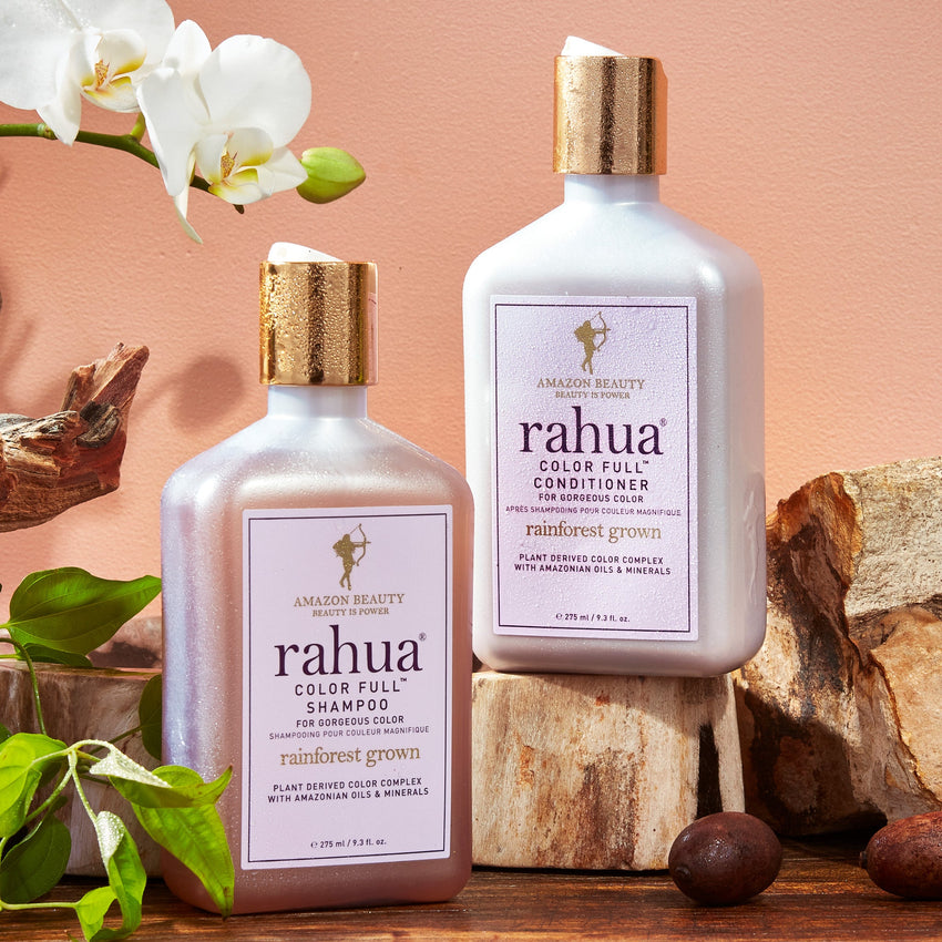 two bottles with gold caps of rahua color full shampoo and conditioner on a plain pink background with gardenia and morete on a wooden slab